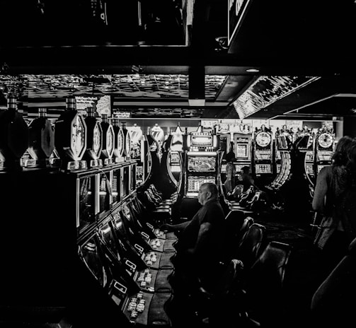 A dimly lit casino filled with rows of slot machines, some occupied by people. The machines' lights contrast with the dark surroundings, creating a vibrant yet subdued atmosphere.