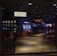 A dimly lit casino with several rows of slot machines, some displaying bright graphics and lights. There are a few people scattered throughout the area, engaging with the machines. The floor is reflective and polished, adding to the ambiance, while the ceiling is dotted with numerous small lights.