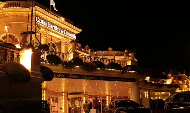 A luxurious casino at night, illuminated with warm, golden lights. The building features elegant architecture with columns and decorative elements. A few people are walking towards the entrance, and there are a couple of parked cars nearby.