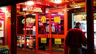 A brightly lit entrance to a casino with neon signs, including one advertising a £500 jackpot. The doors have clear signage indicating entry is restricted to those over 18. A person in a red jacket stands to the right, possibly interacting with a machine or display.