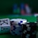 A casino scene featuring two playing cards, the ace and king of clubs, partially concealed by a player's hand. Nearby, there are stacks of multicolored poker chips on a green felt table, with blurred colorful background elements suggesting other chip stacks or cards.