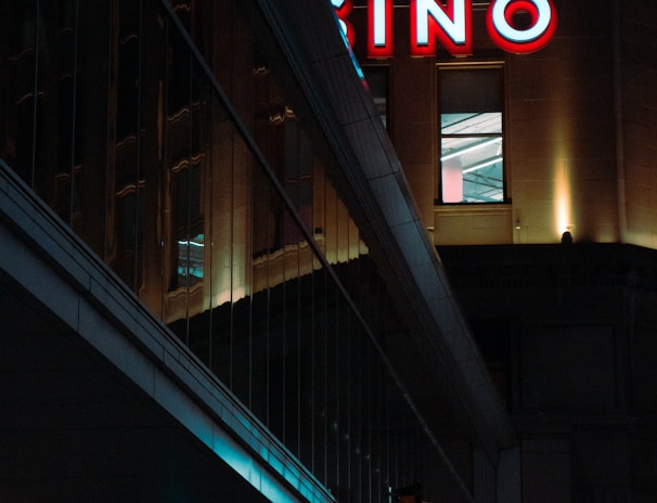 A night scene featuring a building with illuminated neon signage displaying 'CASINO'. The environment is urban with reflections on glass windows and glowing street traffic lights below. The atmosphere is dimly lit, with artificial lighting casting vibrant colors.