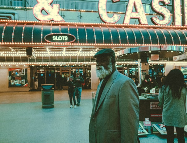 An older man with a gray beard and wearing a suit and hat stands in front of a brightly lit casino. The background features a large illuminated sign reading 'CASINO' and 'SLOTS' among other words, with numerous lights outlining the entrance. Several other people are visible in the background, engaged in various activities, some walking, some standing, and one looking at something on display.