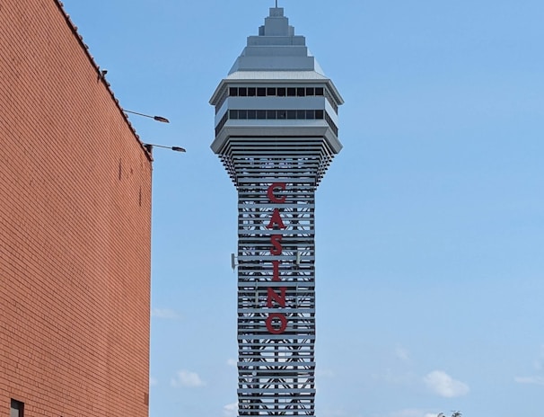 A tall tower with a modern design features the large vertical red letters spelling 'CASINO'. The structure has a sleek, metallic appearance with various horizontal and vertical lines. The sky is mostly clear with a few clouds. In the foreground, part of a brick building is visible, along with some trees and buildings in the distance.